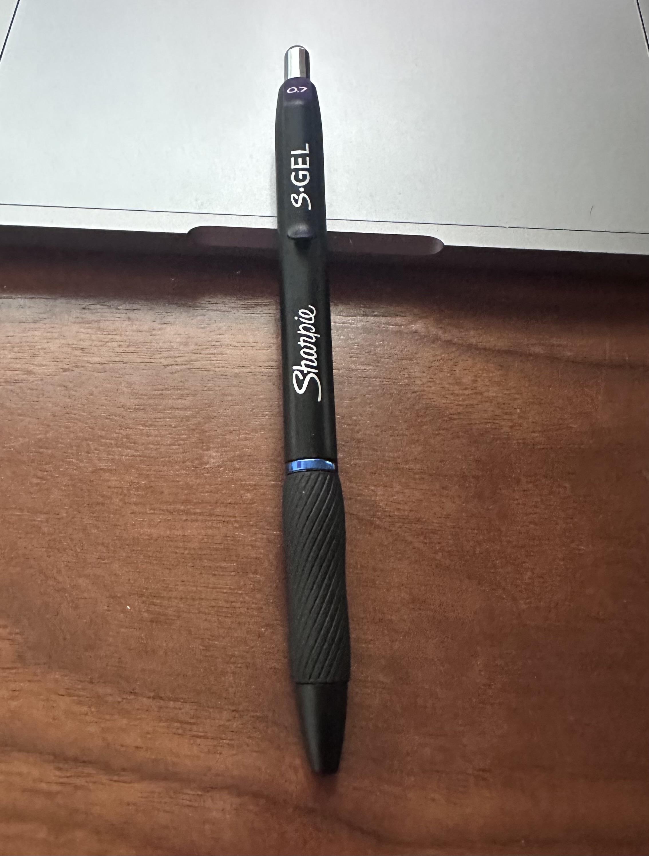 A black Sharpie S-Gel pen with a 0.7mm tip is resting on a wooden surface and laptop. 
