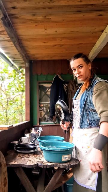 Photograph of a person washing dishes. The person is looking at the camera and holds a pan.