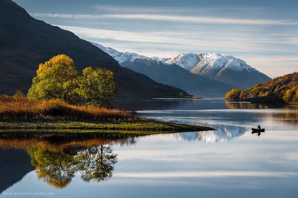 A photograph of a calm Highland lake surrounded by golden autumn trees, their bright leaves mirrored in the still water below. A single canoeist glides quietly across the loch, appearing small against the backdrop of tall, snow-dusted mountains in the distance. Above, a soft morning sky adds to the sense of peace and stillness in the scene.