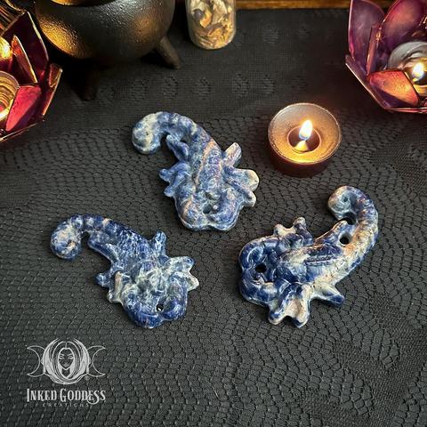 Three specimens of the "Sodalite Large Scorpion" product are arranged on a lace-covered surface. They are in shades of blue and cream and intricately carved into the shape of a resting scorpion. Presented by Inked Goddess Creations.