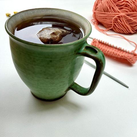 Angled view of a large cup of tea on a white plank background, with a salmon pink ball of yarn, and a knitting needle with a swatch on it in the background.