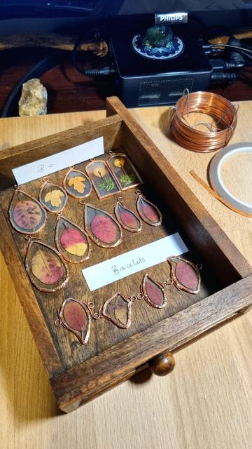 English A wooden drawer with pieces of glass that follow the shape of the plants underneath (autumn leaves and mushrooms), wrapped in copper foil with copper rings above them.
Français Un tirroir en bois avec des morceaux de verre qui épousent la forme des végétaux en-dessous (feuilles d'automne et champignons), entourés d'un ruban de cuivre avec des anneaux en cuivre au-dessus.