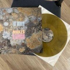 Eine Schallplatte mit einem bunten, strukturierten Cover mit dem Titel "The World Makes A Fool" liegt neben einer transparenten, goldenen Schallplatte auf einer hellen Holzoberfläche.
