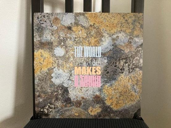 Ein Albumcover mit einem strukturierten, steinähnlichen Hintergrund und einem bunten Text, der lautet: "THE WORLD BARELY MAKES A SOUND". Das Album ist vor einem Holzstuhl ausgestellt.