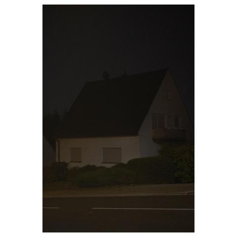 Ein weißes Haus mit schwarzem Dach steht an einer Straße. Es ist Nacht und das Haus nur schwach von einer nicht im Bild sichtbaren Lichtquelle beleuchtet. 