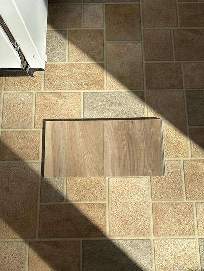 Two samples were f Lifeproof Ledge Point Pecan next to each other, on a sheet vinyl floor that consists of beige and grey faux stones. The LVP pecan samples are a medium brown sandy beige color, with light and dark streaks in the pecan grain. They fall within a big streak of morning sunlight. The corner of a white fridge peaks out of the top left corner.  