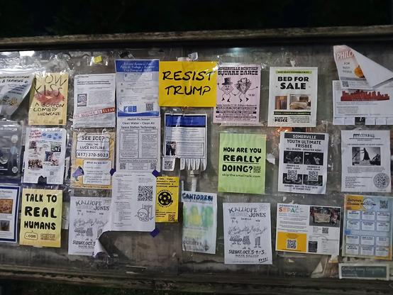 A large community message board with all kinds of messages pinned up. Most are letter sized papers, in pretty even rows and columns (which is unusual).

The center top message printed on bright yellow paper says "RESIST TRUMP".

Other items on the board, which consists of 3 rows and about 8 columns. 
Top row: "Now Kiss" lips and a Boston Comedy showcase, a scientific paper about climate change, job and resource fair in 3 languages, Resist Trump, Square dancing, bed for sale, I think some English tutoring.

Middle row: Language tutoring,  SEE ICE? Call LUCE HOTLINE 617-370-5023, some kind of water and air filtering thing, Polyamory support group, How are you really doing?, youth ultimate frisbee, silent film cinema something.

Lowest row: Talk to real humans, musicians at Nave art gallery, Somerville Cine-club, washed out something I can't read, Nave musicians again--Kalliope Jonesw with Lou, Isabella, and Wes,  Somerville SEPAC special education parents advisory council. 