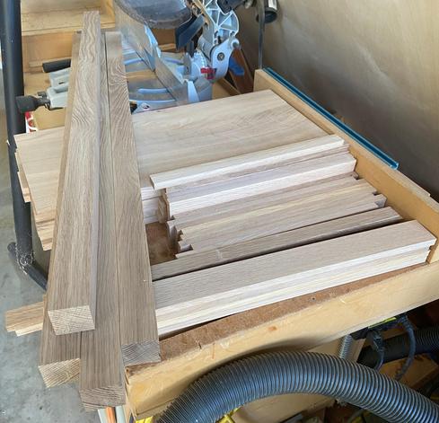 A collection of quarter sawn white oak lumber, cut to width and planed to the correct thickness, sit atop a workbench.  A chop saw is in the background and the hose to a shop vac is in the foreground.