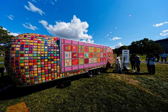 A large trailer has a screen with korean color squares surrounded by little tiny brightly colored squares tiling the rest of it while several students mill around to the right under a blue sky