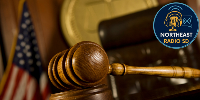 Gavel on desk with blurred American flag in background. "NORTHEAST RADIO SD" logo in corner. Warm, formal courtroom setting.
