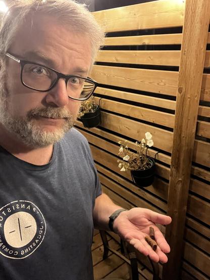 A person with glasses is holding a small object in their hand while standing in front of a wooden slatted wall with hanging flowerpots.