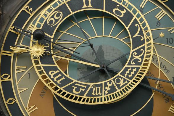 A close-up view of an astronomical clock face featuring ornate gold and black designs with numbers, zodiac symbols, and a sun motif. The clock has intricate hands pointing to various markings.