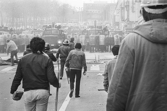 Scène de rue.
Au fond, des policiers de face, avec leurs boucliers.
À l'avant, des ouvriers de dos tenant des barres de fer, pavés 