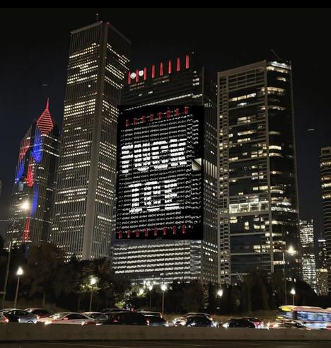 Nighttime cityscape of downtown Chicago with illuminated skyscrapers. The central building displays a bold digital sign reading “FUCK ICE” in white letters on a black background. Surrounding buildings glow with red and blue lights. Busy road in foreground with cars in motion. Image conveys urban protest and anti-ICE sentiment.
