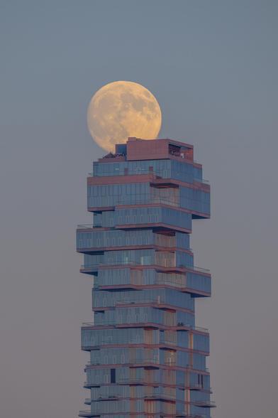 A telephoto snapshot of the almost full moon rising above a conspicuous skyscraper in New York city, taken on October 5, 2025. The 56 Leonard Street skyscraper, is also known as the Jenga Tower, as it indeed is reminiscent of a pile of stacked blocks of a Jenga game.