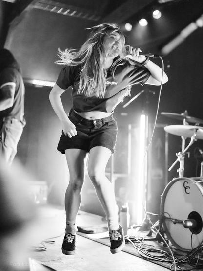 A performer with long hair is singing passionately on stage, jumping in mid-air. They wear a black t-shirt, high-waisted shorts, and sneakers. The scene is set in a dimly lit venue, with a drum kit visible in the background.