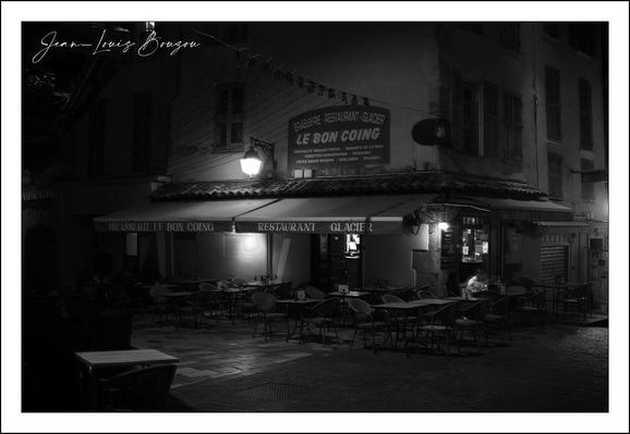 This black and white photograph captures a quiet nighttime scene outside a quaint café or small restaurant named "Le Bon Coing." The establishment is nestled on a corner, with its awning stretching over a collection of empty tables and chairs arranged neatly in front. The soft glow of several wall-mounted lamps casts gentle pools of light, creating an inviting ambiance amid the surrounding darkness.

The building’s textured façade and the cobblestone street contribute to a charming old-world feel, suggesting a Parisian or European setting. The signage above the café declares it a "Restaurant - Glacier," implying it serves both meals and ice cream, blending culinary warmth with casual enjoyment.
The absence of people and the deep shadows lend the image a contemplative, almost melancholic tone—a peaceful pause in the hustle of daily life. The use of monochrome accentuates contrasts and textures, enhancing the timeless, nostalgic essence of the scene.
Artistically, such a photo can symbolize solitude, quiet moments of reflection, or the beauty of simple places that live on quietly after the crowds have gone. It might also evoke the cultural atmosphere of European café culture—an important social and artistic tradition where conversations, thoughts, and ideas once flourished.