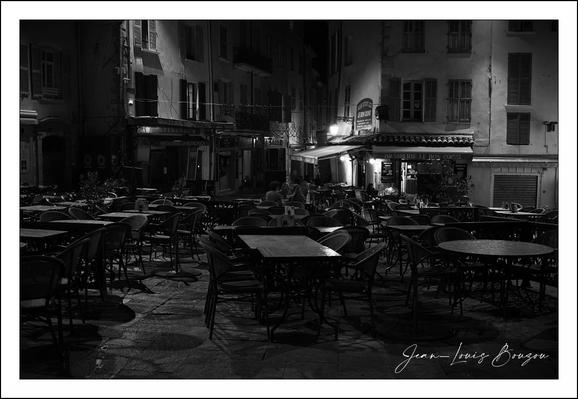 This black-and-white photograph captures the quiet stillness of an empty outdoor café at night. The absence of people, paired with the scattered arrangement of tables and chairs, evokes a sense of solitude and calm. The image plays beautifully with contrasts: the softly illuminated areas from streetlights and building windows sparkle against the deep shadows that cloak parts of the scene, enhancing the timeless atmosphere.
The cobblestone ground and architectural style hint at a historic European setting, with tall, closely packed buildings lining the back of the café. The light source seems almost theatrical, spotlighting certain tables and parts of the empty chairs, drawing attention to the textures of the stone and wrought iron.
Artistically, the photograph might symbolize moments of pause and reflection amid the usual bustle of life. The emptiness can speak to themes of absence, waiting, or the end of a day, inviting the viewer to contemplate stories that unfold in the quiet hours. The monochrome palette strips away distractions, focusing the eye on form and shadow, adding a layer of nostalgia and introspection.
Overall, the image is a poetic snapshot of urban life frozen in a tranquil moment, its silent streets telling a story without words.
