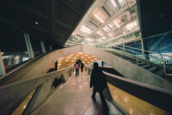 Several people who are facing awat from the camera and disrorted are walking away on a birdge on a concrete bridge that goes under a shell connected to by other bridges with staircases,