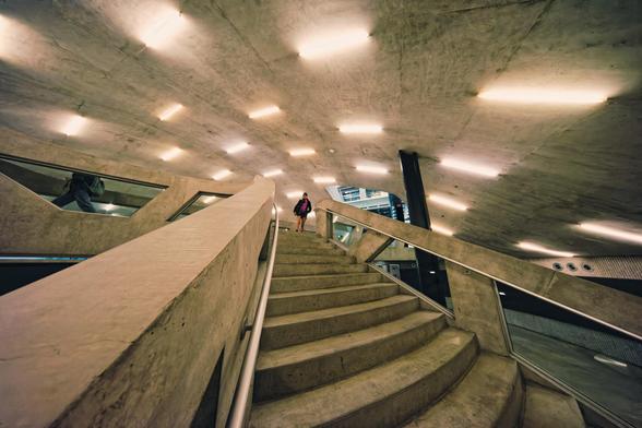 A woman is indistinct at the top of a concrete stair with coarse railings lit by a number of linear fixtures laid out on a hyperbolic shell.
