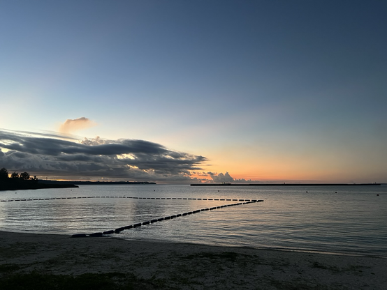 A sandy beach runs to the ocean where in the background you can see the sun setting behind the clouds. A swimming area is marked in the water 