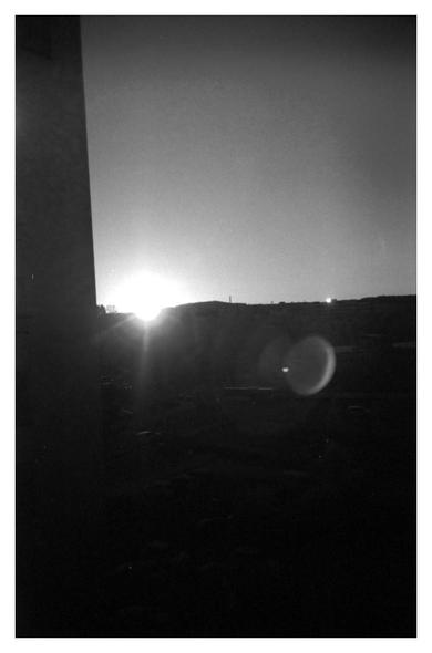 A black and white photo of a sunrise. Cloudless sky, the sun has just crept over the distant landskape causing a flate in the lens. A tall building is partially blocking the view to the left.
