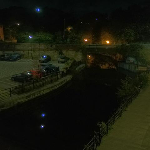 Photograph of the tidal stretch of Ouseburn River at south side of Ouseburn Bridge in Newcastle upon Tyne. To the left (south-west) of the bridge is Cut Bank, and to the right (north-east) is Byker Bank. In the foreground is the pedestrian Riverside Walk and the photo also spans some surrounding built-up areas and trees, with sky in the background. Still image captured from webcam at 05:52 hrs on Friday 10 October 2025.