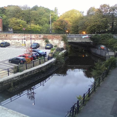 Photograph of the tidal stretch of Ouseburn River at south side of Ouseburn Bridge in Newcastle upon Tyne. To the left (south-west) of the bridge is Cut Bank, and to the right (north-east) is Byker Bank. In the foreground is the pedestrian Riverside Walk and the photo also spans some surrounding built-up areas and trees, with sky in the background. Still image captured from webcam at 07:25 hrs on Friday 10 October 2025.