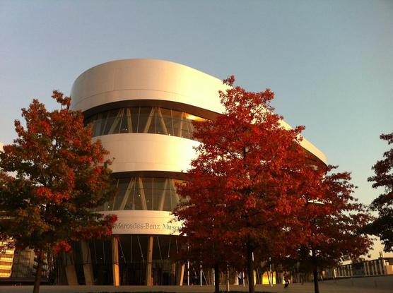 Rounded building with aluminum facade and glass. In front of the building, maple trees with red autumn leaves.