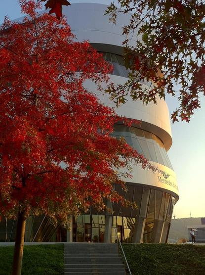 Rounded building with aluminum facade and glass. In front of the building, maple trees with red autumn leaves.