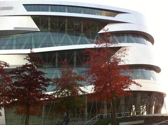 Rounded building with aluminum facade and glass. In front of the building, maple trees with red autumn leaves.