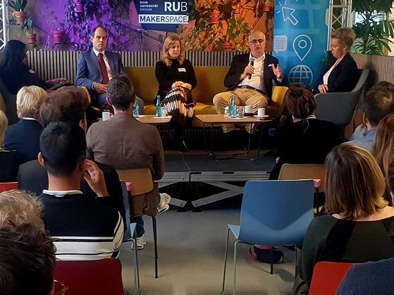 Eine Gruppe von Menschen sitzt auf einer Bühne, an dem ein Mikrofon steht. Der Tisch ist in einem Raum mit roten Stühlen und einer blauen Wand aufgestellt. Auf der Rückwand befindet sich ein Schild mit der Aufschrift „RUB“ und darunter „UNIVERSITÄT BOCHUM“. Darunter befindet sich die Aufschrift „MAKERSPACE“. Einige der Menschen sitzen auf roten Stühlen, andere stehen oder sitzen auf blauen Stühlen. Ein hellgrünes Banner ist an der rechten Seite des Bildes zu sehen.