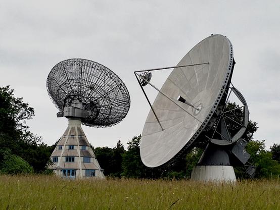 Eine große Antenne in Form einer Parabolschüssel steht auf einem Gebäude in Form eines Kegels. Davor, in einer Wiese, ist eine weitere Parabolantenne zu sehen.