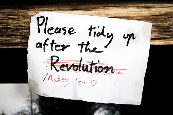 A small scrap of paper is attached to a solid timber bench, bearing the handwritten phrase, "Please tidy up after the Revolution". The word "Revolution" is scribbled out in finer red pen, and the phrase "Making Tea <3" appended.