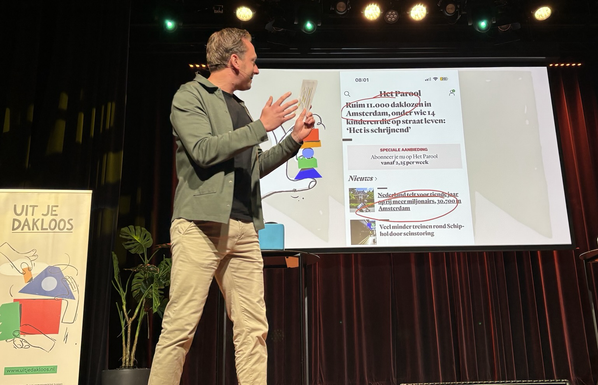 Foto van een podium vanaf de eerste rij in de zaal genomen. De presentator wijst naar een scherm met screenshots van krantenkoppen over dakloosheid en de groei van het aantal miljonairs. Links de banner van Uit je Dakloos.