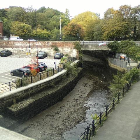 Photograph of the tidal stretch of Ouseburn River at south side of Ouseburn Bridge in Newcastle upon Tyne. To the left (south-west) of the bridge is Cut Bank, and to the right (north-east) is Byker Bank. In the foreground is the pedestrian Riverside Walk and the photo also spans some surrounding built-up areas and trees, with sky in the background. Still image captured from webcam at 12:28 hrs on Friday 10 October 2025.
