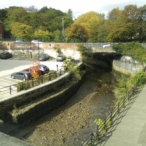 Photograph of the tidal stretch of Ouseburn River at south side of Ouseburn Bridge in Newcastle upon Tyne. To the left (south-west) of the bridge is Cut Bank, and to the right (north-east) is Byker Bank. In the foreground is the pedestrian Riverside Walk and the photo also spans some surrounding built-up areas and trees, with sky in the background. Still image captured from webcam at 12:44 hrs on Friday 10 October 2025.