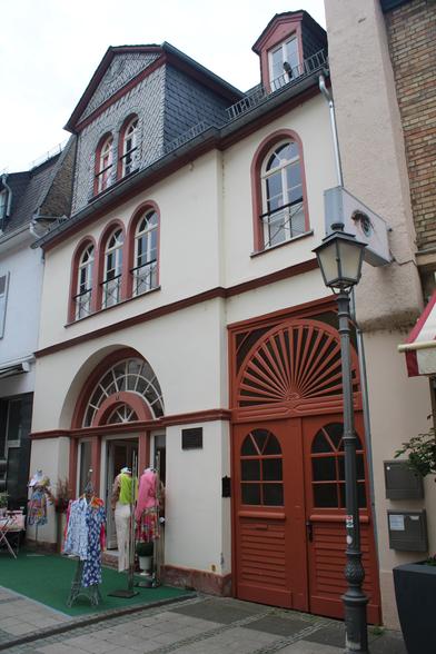 Ein schräger Blick auf ein dreigeschossiges Gebäude. Das Erdgeschoss verfügt über zwei hohe Türen mit Rundbogen. Eine Tür ist geöffnet, davor stehen auf einem Stück Kunstrasen ein paar Ständer mit Kleidung. Das andere Tor ist verschlossen. Die Fassade ist hell-beige und die Fenster weisen ebenfalls rot-brauen Rundbögen auf. Das Dachgeschoss ist mit Schieferschindeln bedeckt. 
