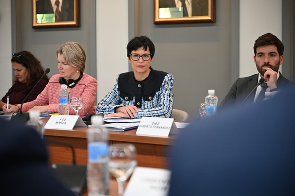Image of four people sitting behind desk with nametags. The woman in the middle with black short hair and glasses looks towards the camera whilst the two women to her left look down at the desk and the man on her right looks ahead with his hand up to this face