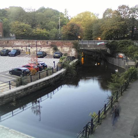 Photograph of the tidal stretch of Ouseburn River at south side of Ouseburn Bridge in Newcastle upon Tyne. To the left (south-west) of the bridge is Cut Bank, and to the right (north-east) is Byker Bank. In the foreground is the pedestrian Riverside Walk and the photo also spans some surrounding built-up areas and trees, with sky in the background. Still image captured from webcam at 18:16 hrs on Friday 10 October 2025.