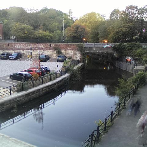 Photograph of the tidal stretch of Ouseburn River at south side of Ouseburn Bridge in Newcastle upon Tyne. To the left (south-west) of the bridge is Cut Bank, and to the right (north-east) is Byker Bank. In the foreground is the pedestrian Riverside Walk and the photo also spans some surrounding built-up areas and trees, with sky in the background. Still image captured from webcam at 18:21 hrs on Friday 10 October 2025.