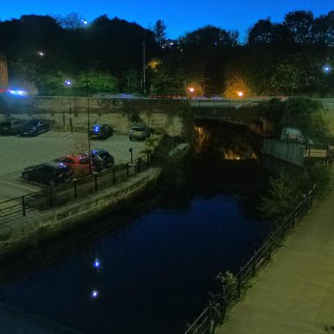Photograph of the tidal stretch of Ouseburn River at south side of Ouseburn Bridge in Newcastle upon Tyne. To the left (south-west) of the bridge is Cut Bank, and to the right (north-east) is Byker Bank. In the foreground is the pedestrian Riverside Walk and the photo also spans some surrounding built-up areas and trees, with sky in the background. Still image captured from webcam at 19:04 hrs on Friday 10 October 2025.