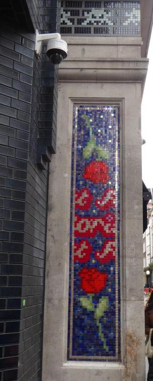 Invader artwork of three invaders, jade on black, above blue and red mosaic of roses on side of building. Security camera top left.