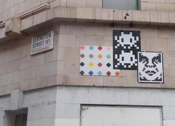 Three Invader artworks on side of building - mosaic of Damien Hirst's ink spots, two white invaders above one another on black mosaic, black and white mosaic face. Sign for Lexington Street to the left. Day.