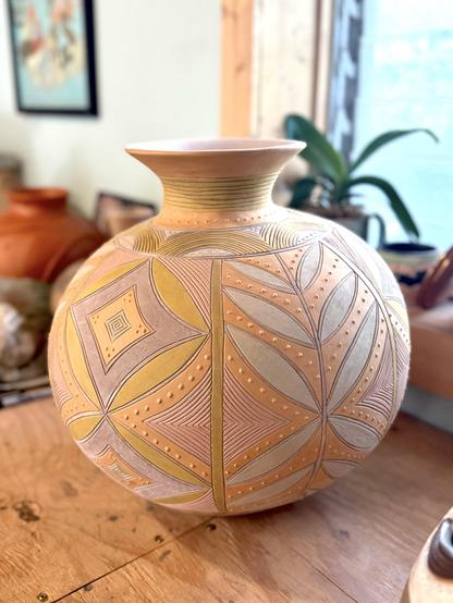 A decorative unfired pottery vase with a spherical body and a narrow neck, featuring intricate geometric and leaf patterns in earthy tones of orange, yellow, and beige. It rests on a wooden surface, with other pottery and plants blurred in the background.