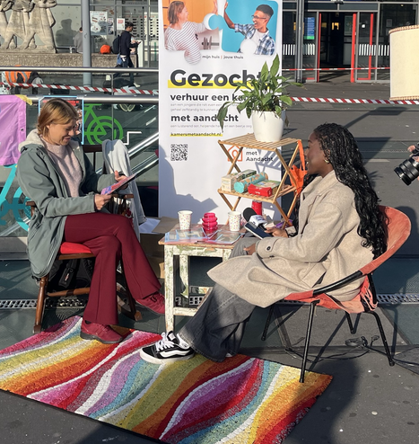 Foto van mij op een stoel aan een tafeltje, buiten bij station Nijmegen. Foto is genomen op het stationsplein. Ik praat met een jonge vrouw die zo een interview gaat afnemen en tegenover me zit. Achter ons een banner van kamers met aandacht.
