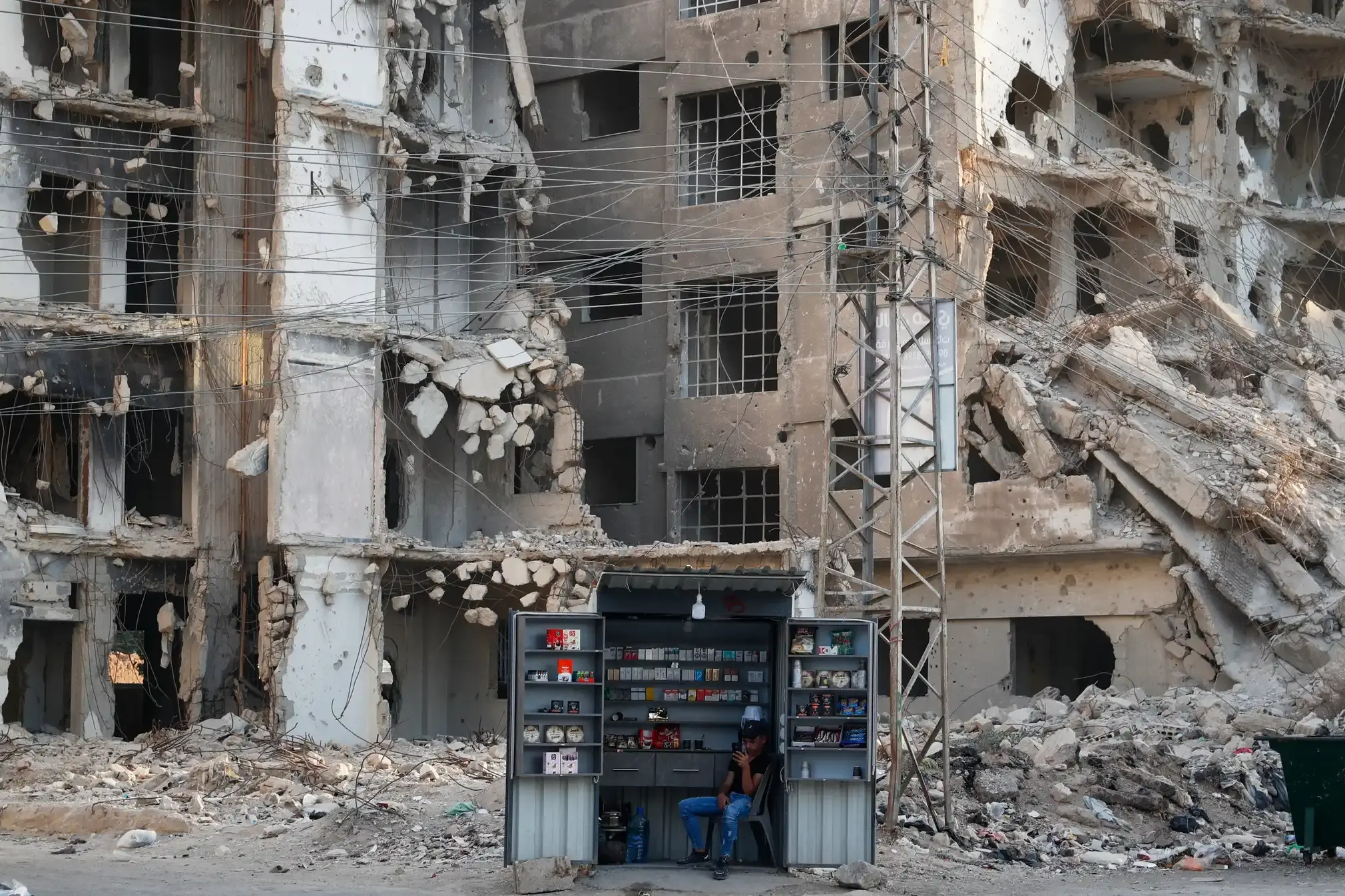 A man waits for customers at a stall in front of war-damaged buildings.