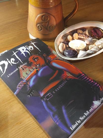 A copy of the short story anthology 'Diet Riot: A Fatterpunk Anthology' (edited by Nico Bell and Sonora Taylor). The cover features an illustration of a fat woman in punk clothing, holding a baseball bat over one shoulder. Next to the book is a large shell with a lit tea light and various bits of plants, bones, and snail shells in it. Above those is a brown ceramic mug with a triple spiral design on the front.