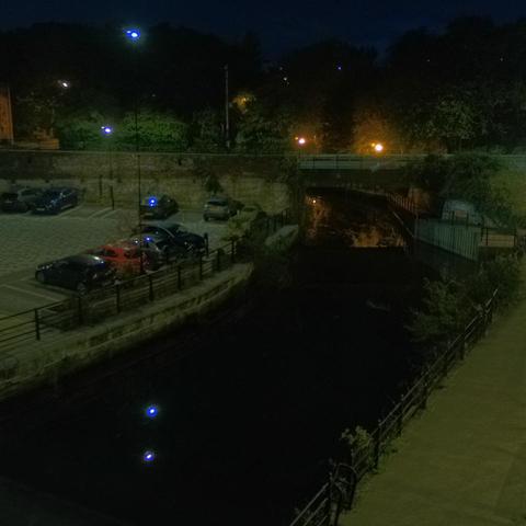 Photograph of the tidal stretch of Ouseburn River at south side of Ouseburn Bridge in Newcastle upon Tyne. To the left (south-west) of the bridge is Cut Bank, and to the right (north-east) is Byker Bank. In the foreground is the pedestrian Riverside Walk and the photo also spans some surrounding built-up areas and trees, with sky in the background. Still image captured from webcam at 06:40 hrs on Saturday 11 October 2025.