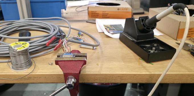 Sarjakaapelin tinaamista kolvilla. Liitin pienessä ruuvipenkissä. Kaapeli kiepillä. Tinaa rullassa. Joitakin työkaluja pöydällä.